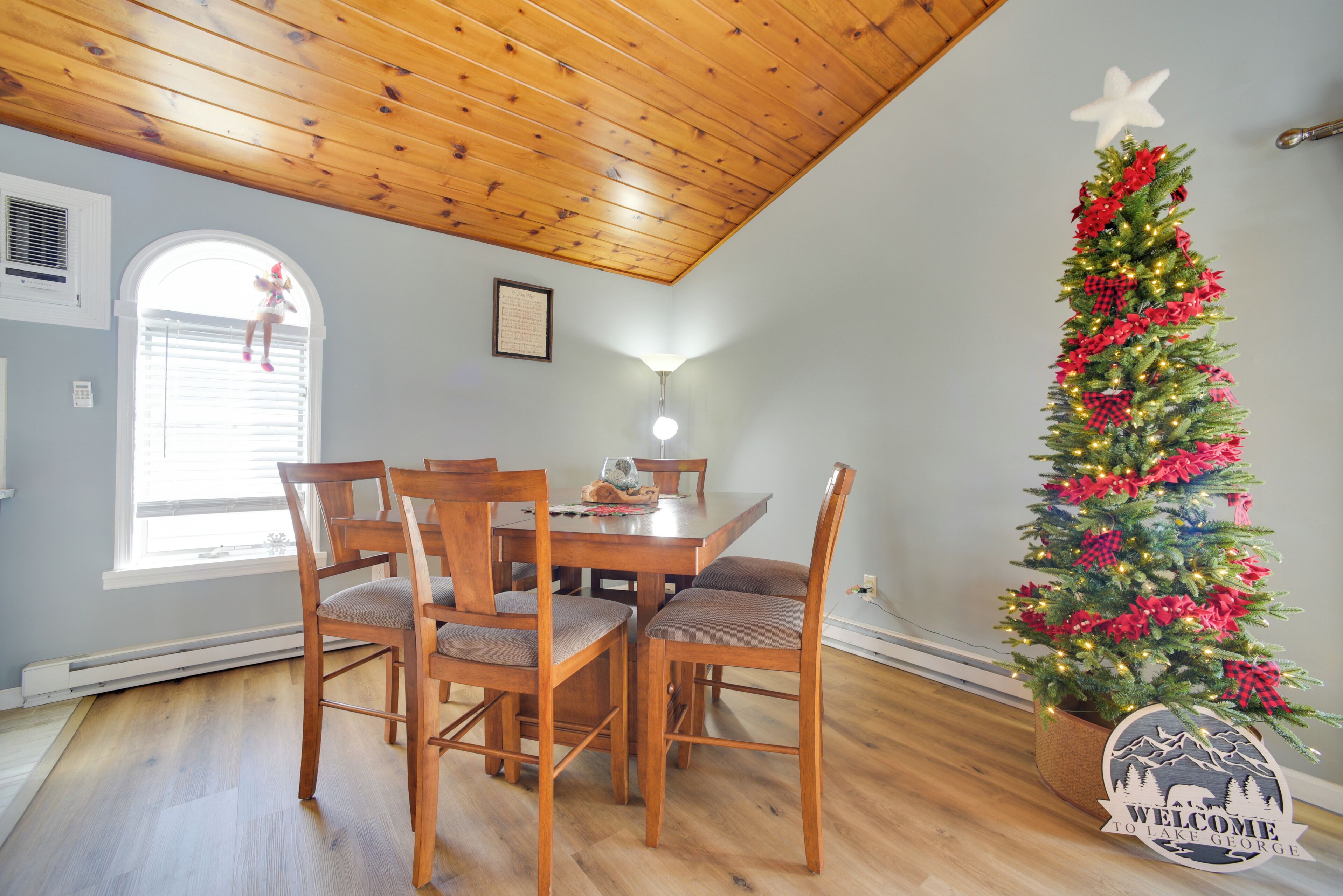 Uma área de refeições com uma mesa e cadeiras de madeira, uma árvore de Natal com luzes e enfeites, e um letreiro com a mensagem 'Welcome to Lake George'.