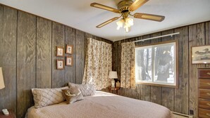 Ein Schlafzimmer mit einer holzverkleideten Wand, einem Bett mit gemustertem Überwurf, einem Deckenventilator und einem Fenster mit Vorhängen.