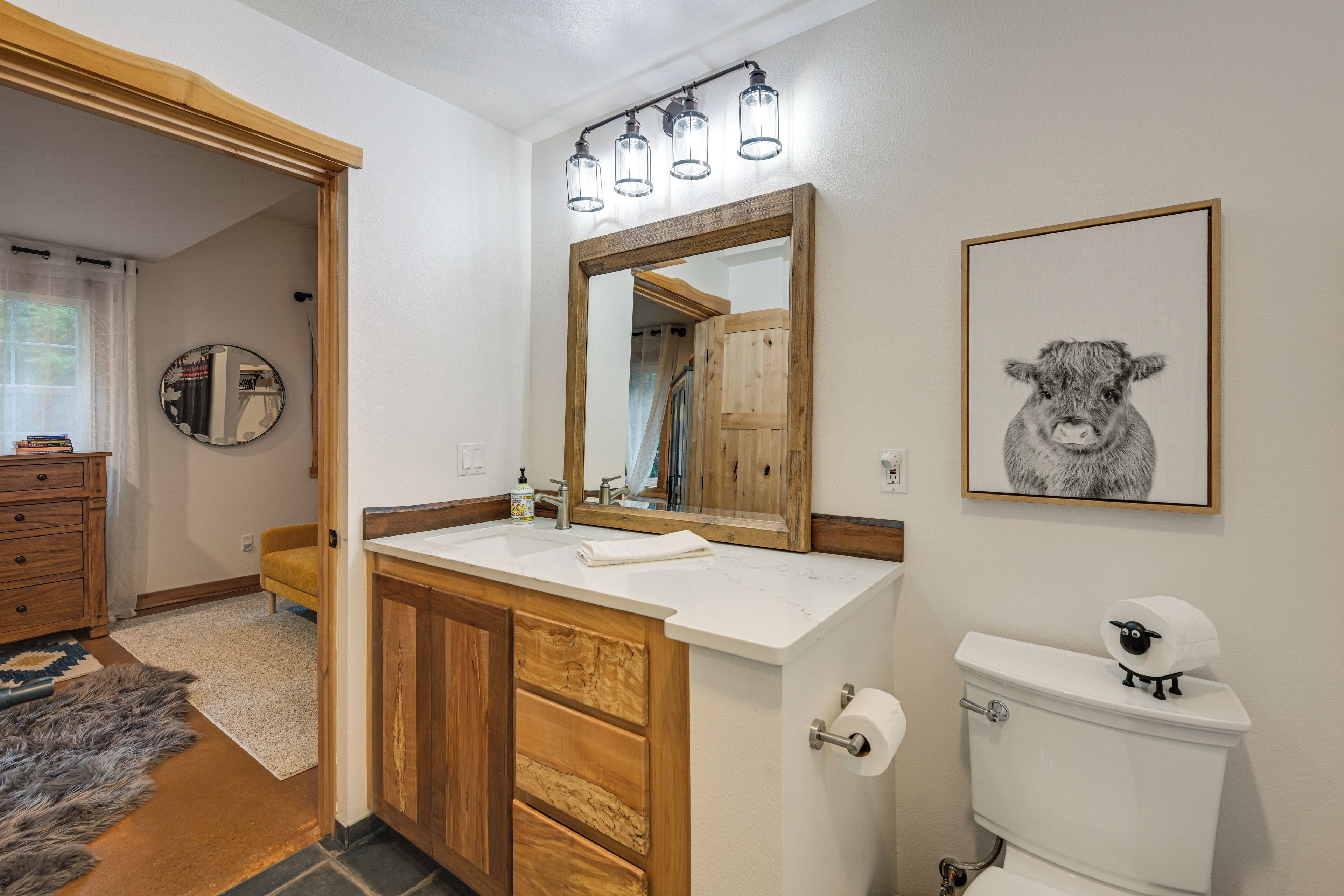 A bathroom with a wooden vanity, a large mirror, a toilet with a sheep decoration, and a framed picture on the wall.