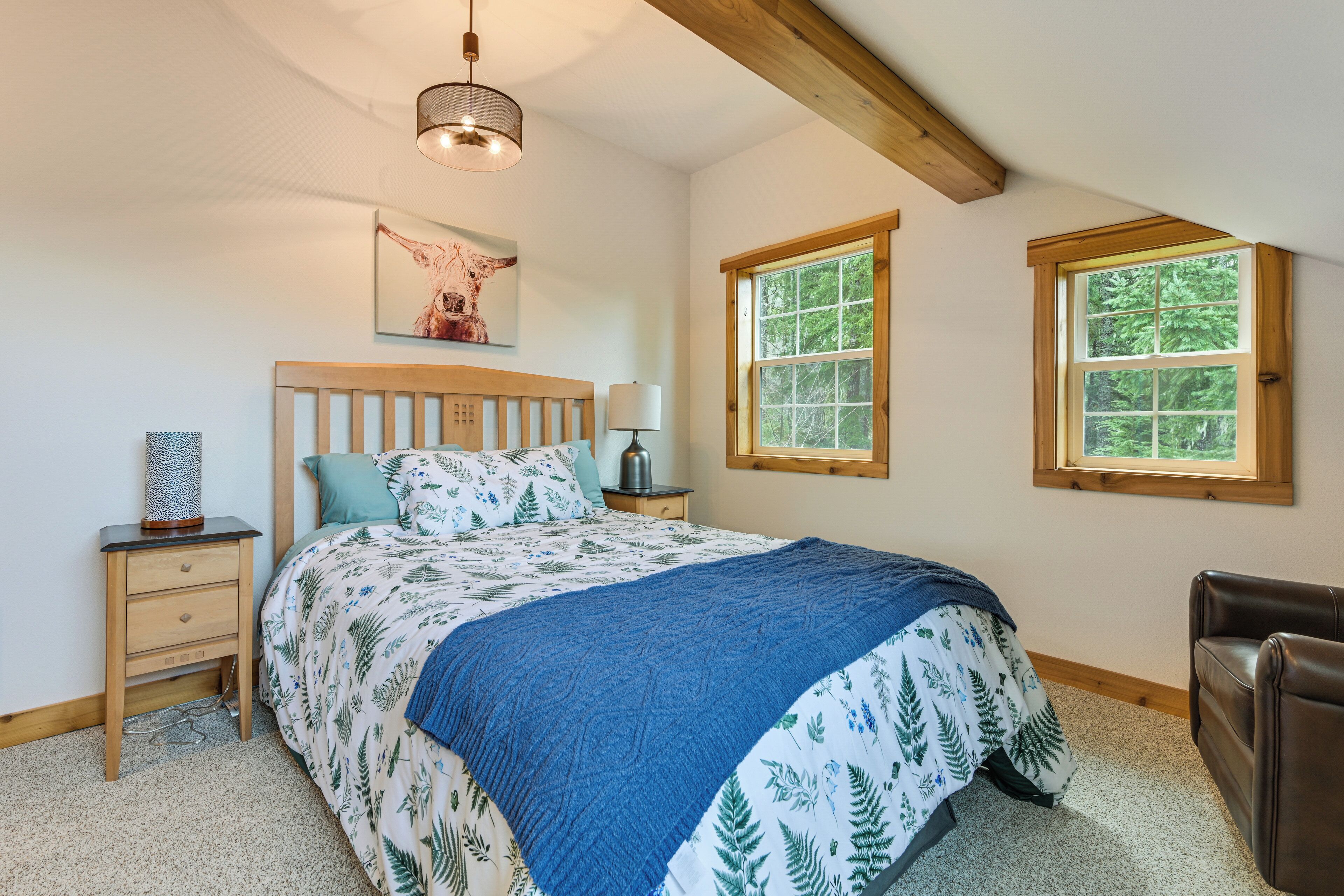 Une chambre à coucher avec une tête de lit en bois, un lit recouvert d’une couvre-lit à motifs floraux, deux fenêtres, une lampe de chevet et un tableau encadré accroché au mur.