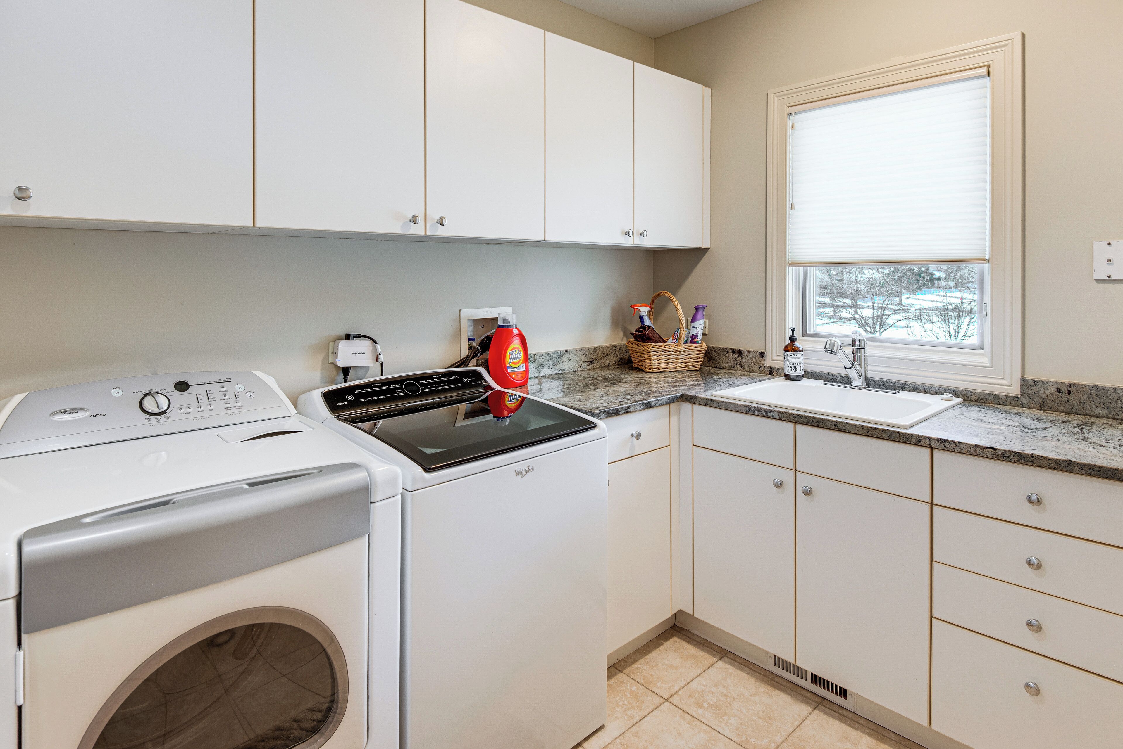 A laundry area with a front-loading washer and dryer, a sink, and a window with blinds.
