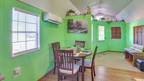 A dining area with a round table, wooden chairs, and a bench. There is a painting on the wall, a window with blinds, and an air conditioning unit. - Cozy Cabin in Overton Glamping Community w/ Perks! (Overton)