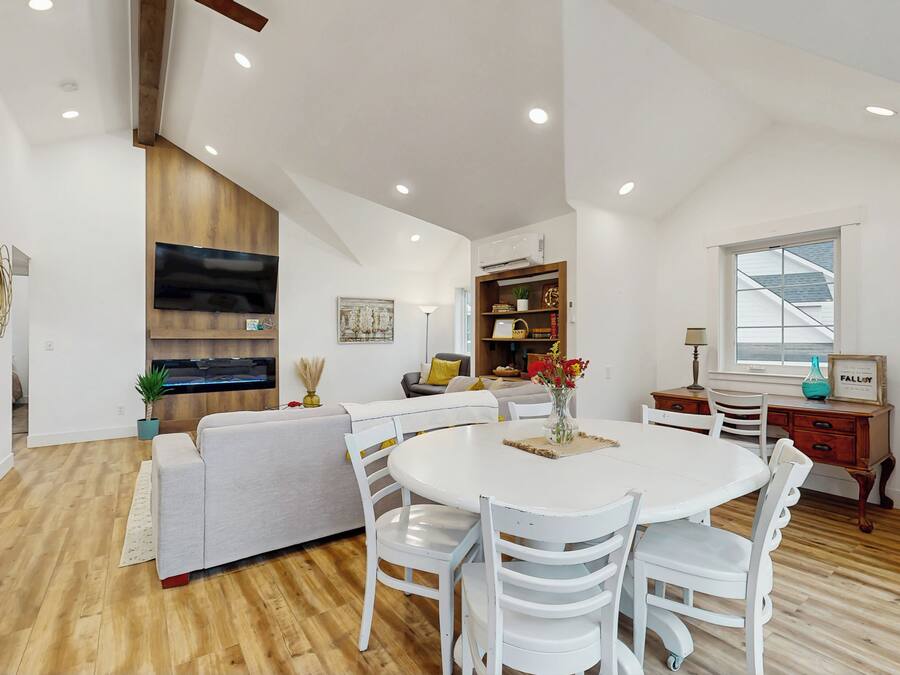 A modern living room with a white sofa, a dining table with white chairs, a flat-screen TV mounted on the wall, and a wooden ceiling.