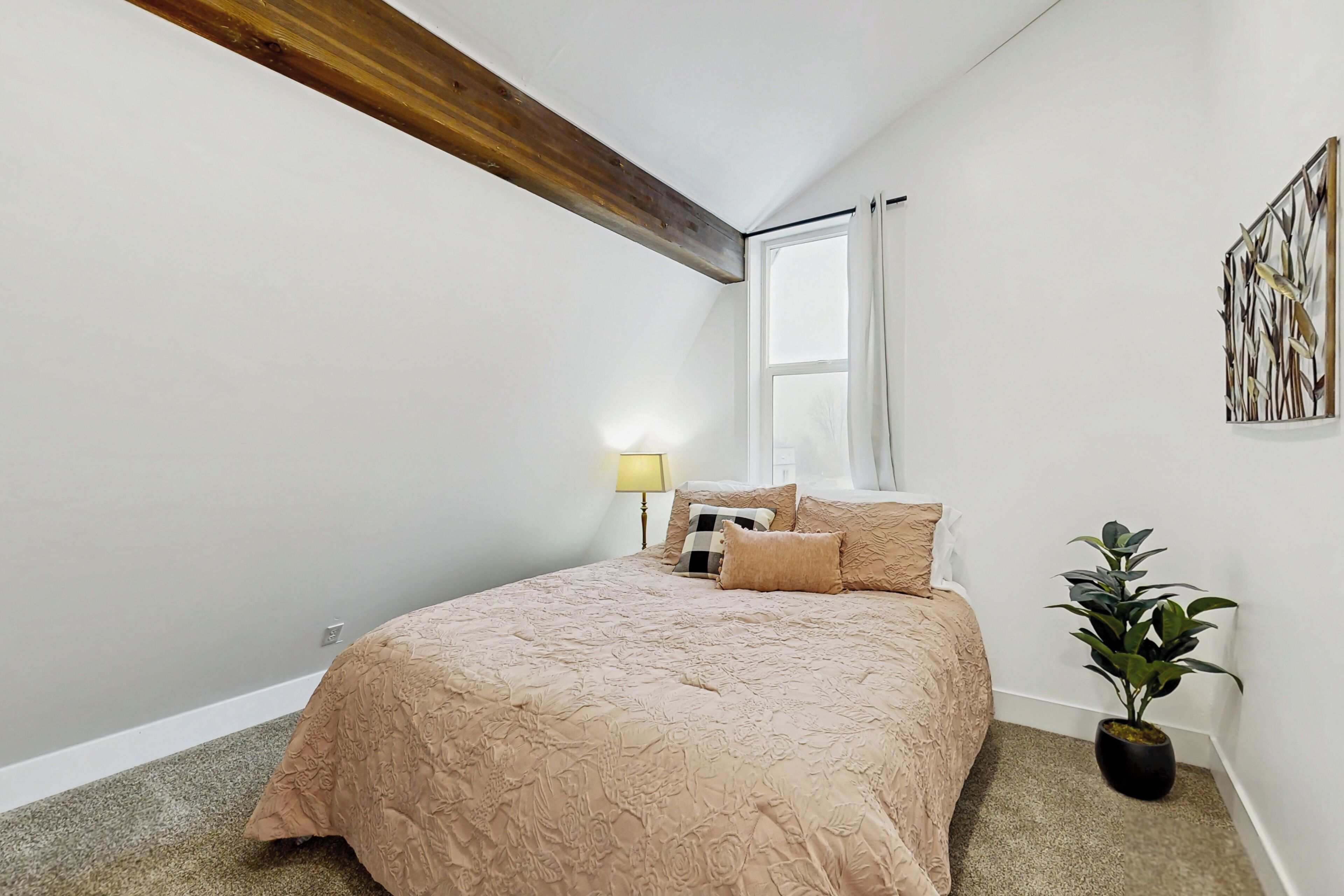 A bedroom with a large bed, a nightstand, a lamp, a window, and a plant.