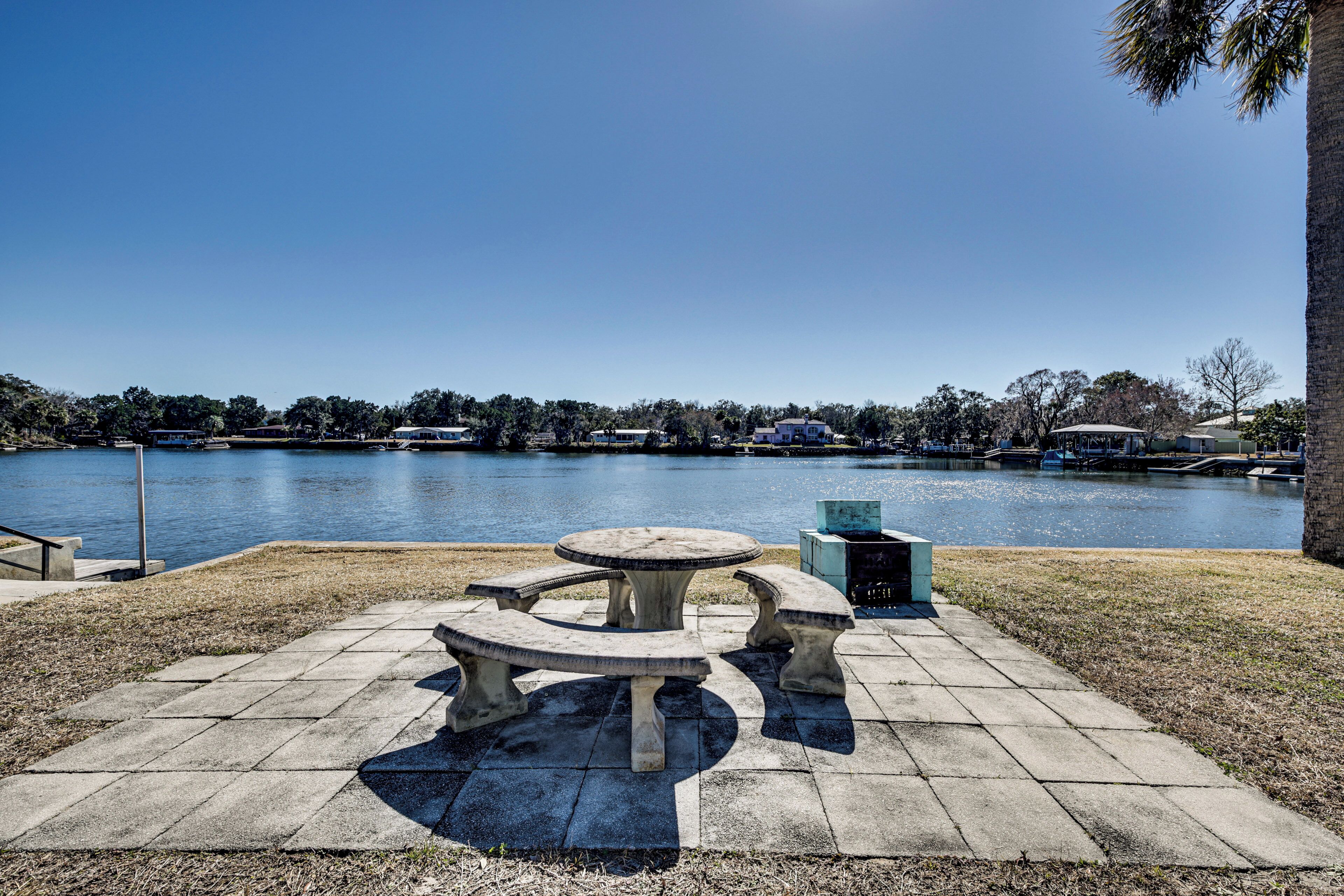A concrete picnic table with benches by a lake, surrounded by grass and a paved walkway.