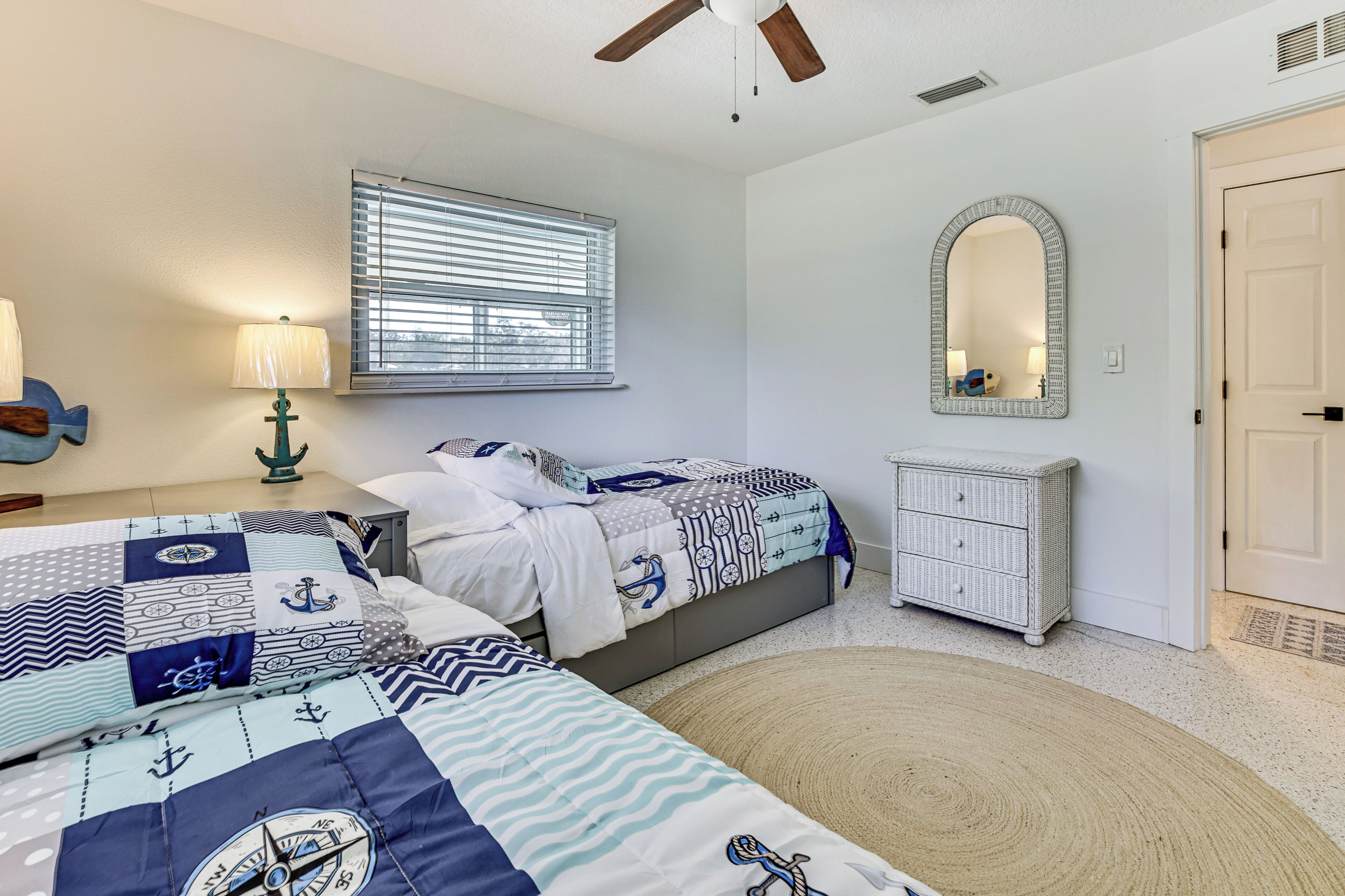 A bedroom with two beds, a window with blinds, a mirror, a dresser, and a ceiling fan.