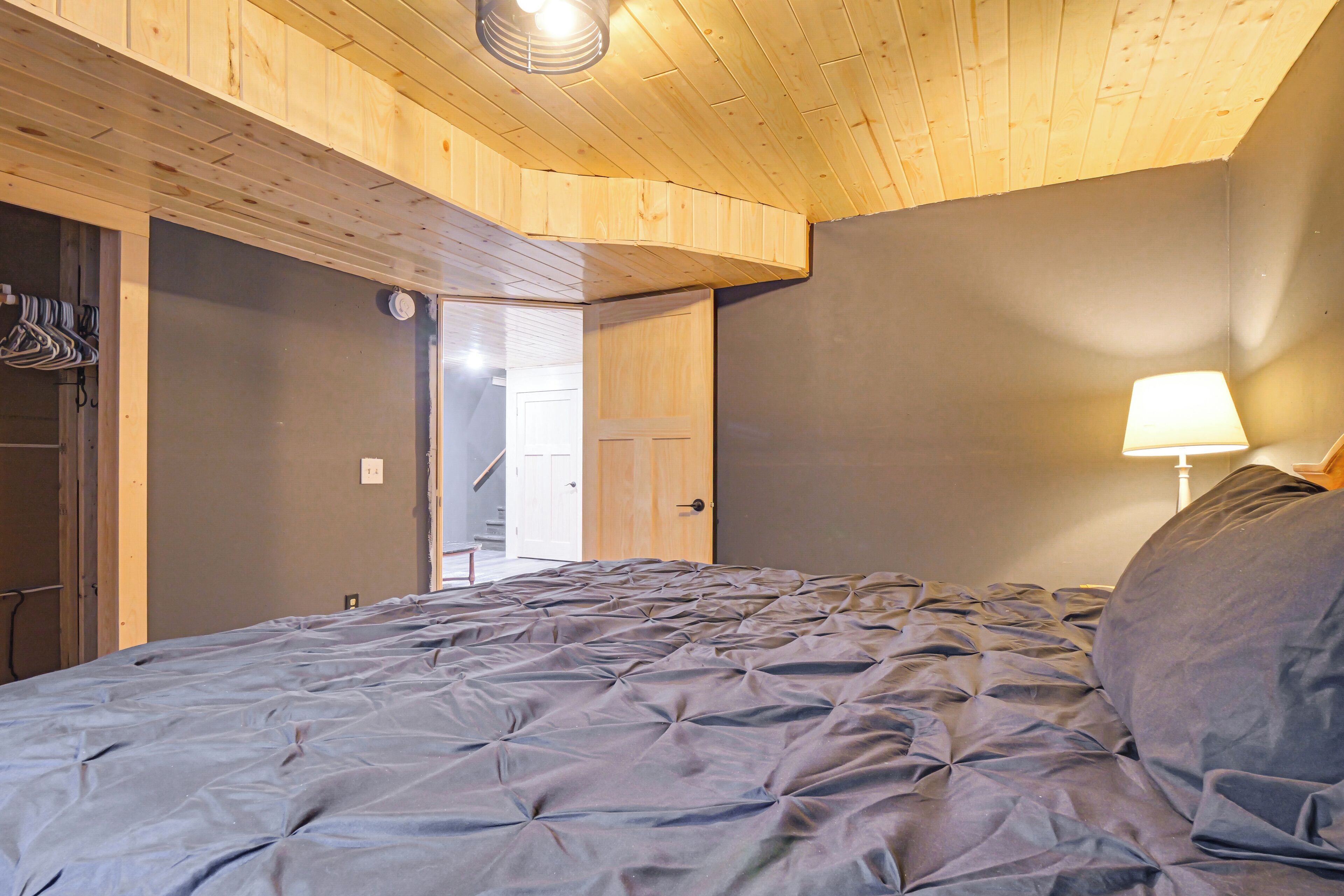 A bedroom with a bed, a wooden ceiling, a door, and a wall-mounted lamp.