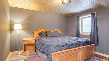 A bedroom with a wooden bed, a nightstand, a lamp, a window with curtains, and a wooden ceiling.