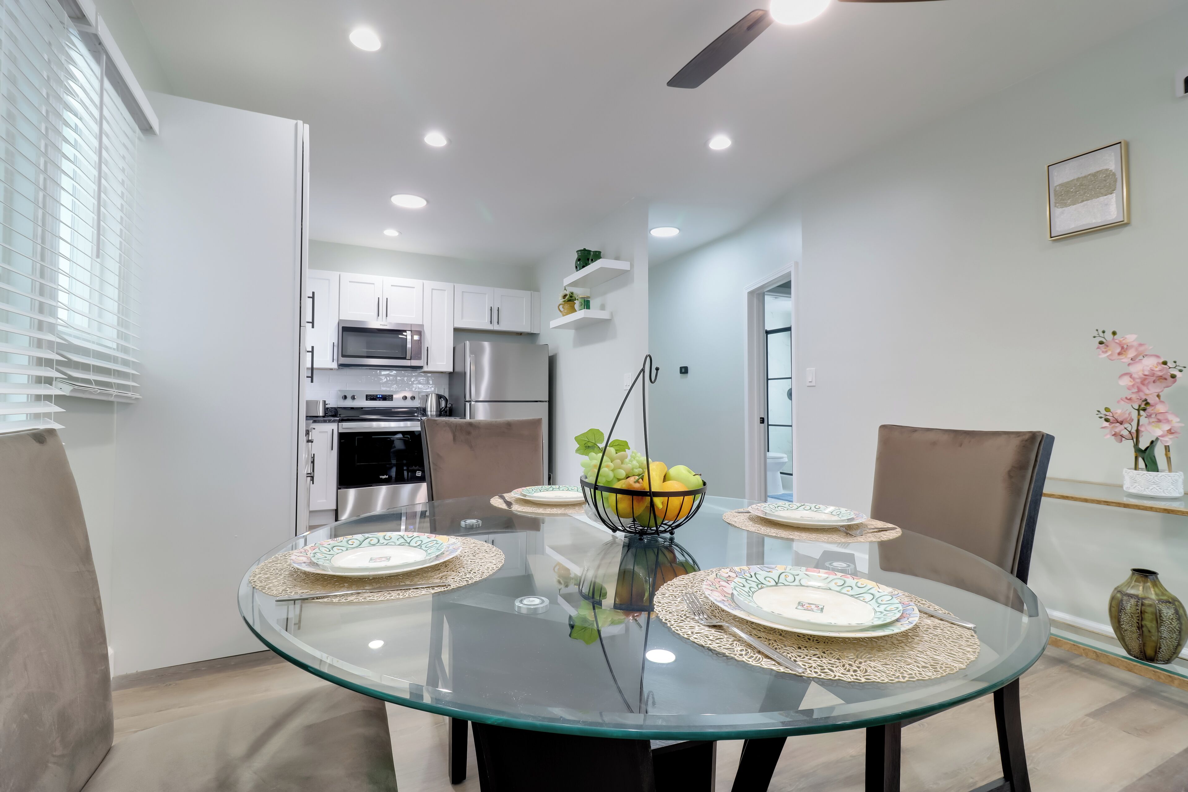 Una cocina moderna con armarios blancos, electrodomésticos de acero inoxidable y un comedor con una mesa de vidrio dispuesta para cuatro.