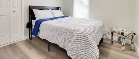 A bedroom with a bed, a white door, a window with blinds, and a small bench.
