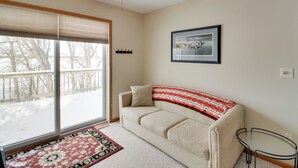 A living room with a beige sofa, a glass coffee table, a ceiling fan, a framed picture on the wall, and a view of a snowy deck through a sliding glass door. - Family Nature Haven! Lakefront Pelican Lake Duplex (Ashby)
