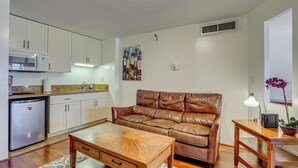 A compact living space with a kitchenette, a brown leather sofa, a wooden coffee table, and a small side table with a lamp and a plant. - Harbor View + Pool Access: Downtown Honolulu Gem! (Honolulu)
