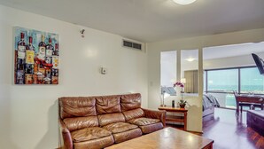 A living room with a brown leather sofa, a wooden coffee table, and a painting of wine bottles on the wall. - Harbor View + Pool Access: Downtown Honolulu Gem! (Honolulu)