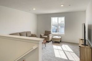 Een ruime woonkamer met een groot raam, een flatscreen televisie op een houten standaard en een beige bank.