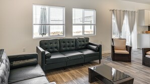 A living room with a dark green sofa, a glass coffee table, and a wicker chair. There are two windows with blinds and a curtain. - Pet-friendly Yard! Family Home in Bakersfield (Bakersfield)