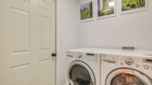 A laundry room with a front-loading washing machine and dryer, a white door, and three framed pictures on the wall. - Mcm-inspired Decor & Top Location! Tumwater Duplex (Olympia)