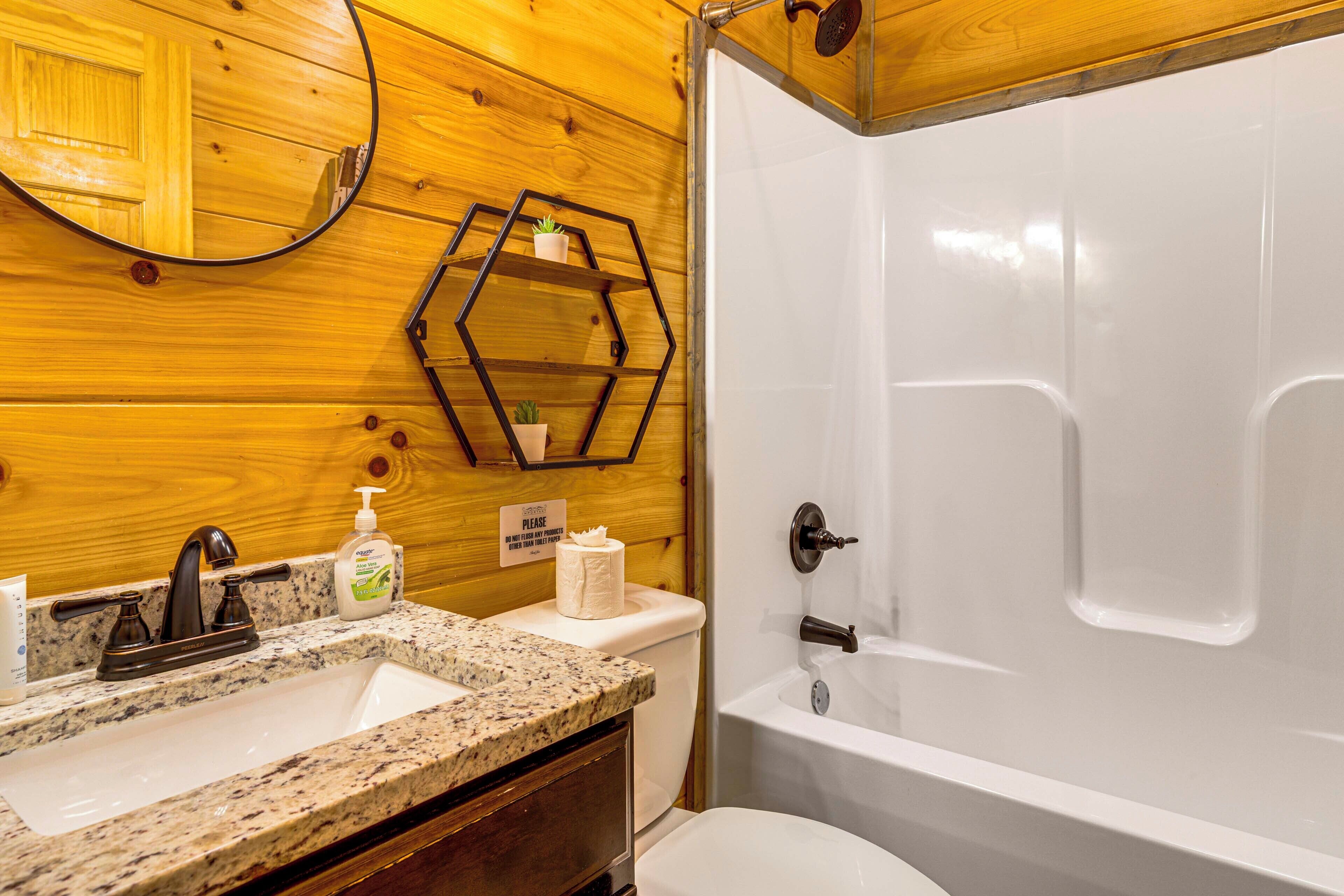A bathroom with a granite countertop sink, a toilet, a bathtub, and a wooden wall with a mirror and hexagonal shelves.