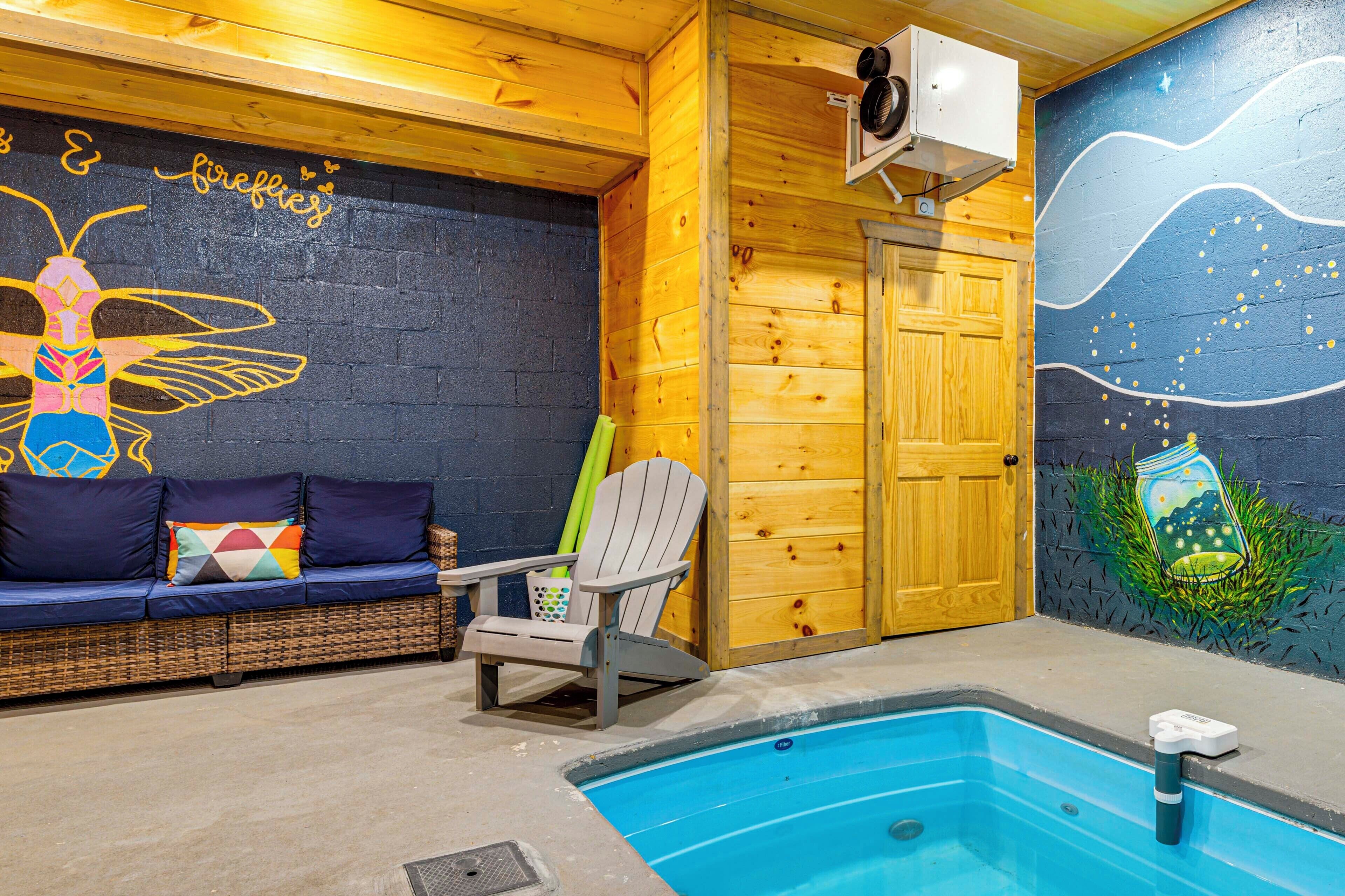 Un área de piscina interior acogedora con una pared de madera, un mural, un sofá con cojines, una silla y una piscina con un flotador.