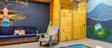 Un área de piscina interior acogedora con una pared de madera, un mural, un sofá con cojines, una silla y una piscina con un flotador.