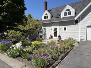 A two-story house with a front porch, a chimney, and a garden with various plants and flowers.