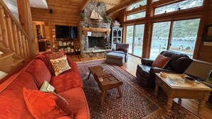 A cozy log cabin interior with a stone fireplace, a television, and a large window overlooking a snowy landscape.