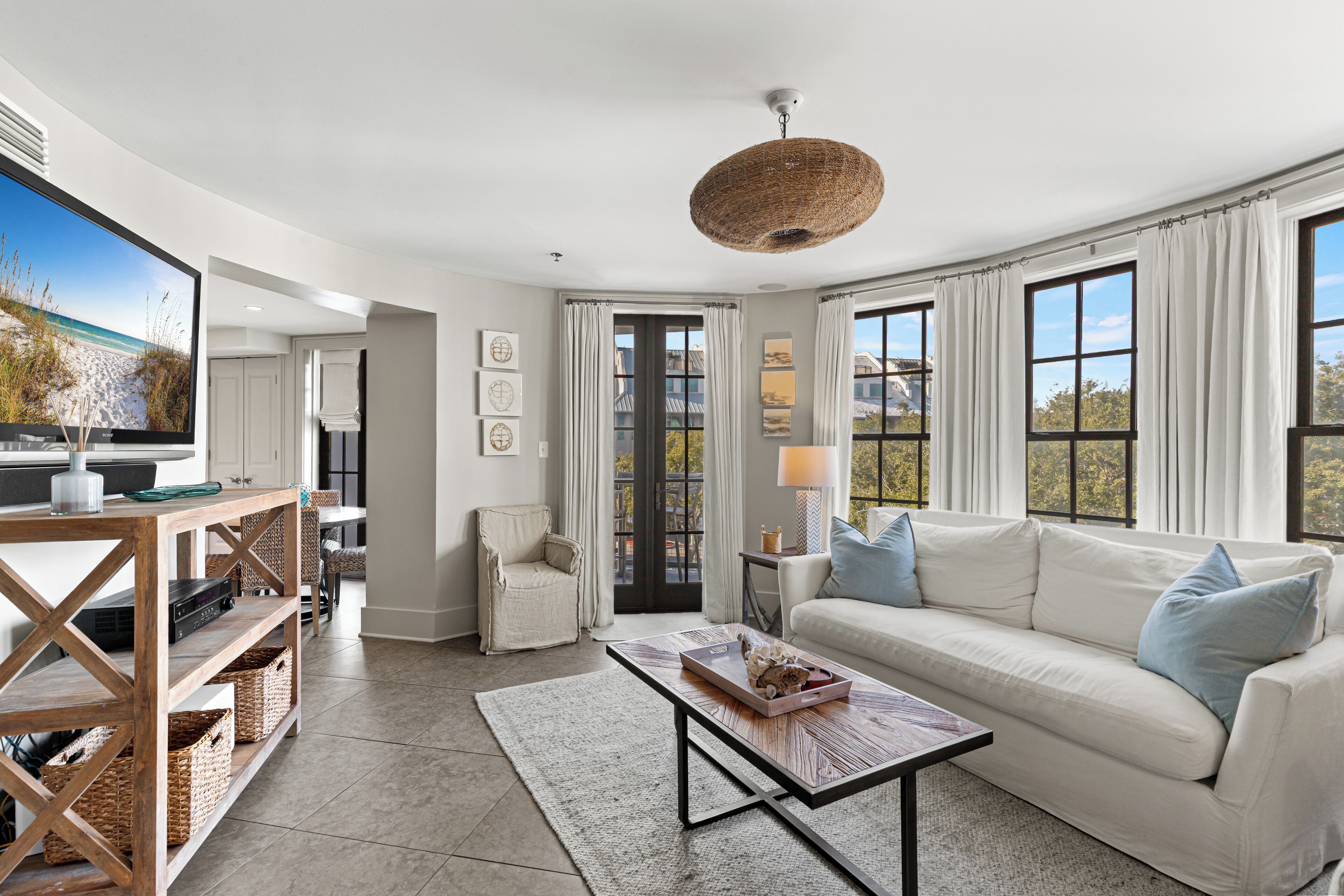 A modern living room with a white sofa, a coffee table, and a TV on a wooden stand. There are large windows with curtains, a wicker ceiling light, and a view of the outdoors.