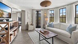 Ein modernes Wohnzimmer mit einem weißen Sofa, einem Couchtisch und einem Fernseher auf einem Holzboden. Große Fenster mit Vorhängen, eine Korbhängeleuchte und ein Blick ins Freie.