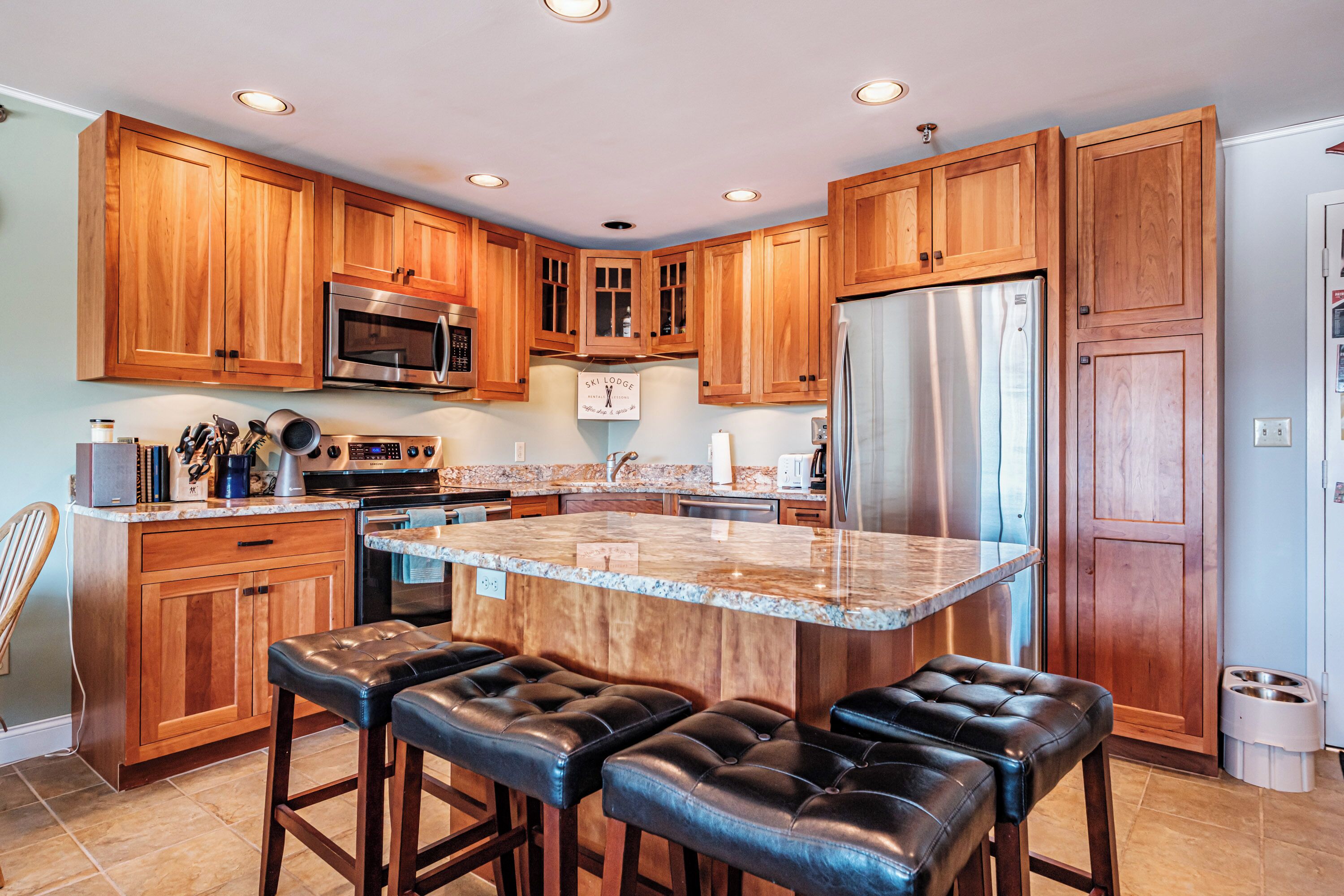 A kitchen with wooden cabinets, stainless steel appliances, a marble island with bar stools, and a dog feeding station.