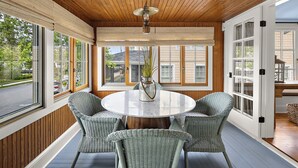 A room with a round table, wicker chairs, a potted plant, and large windows.
