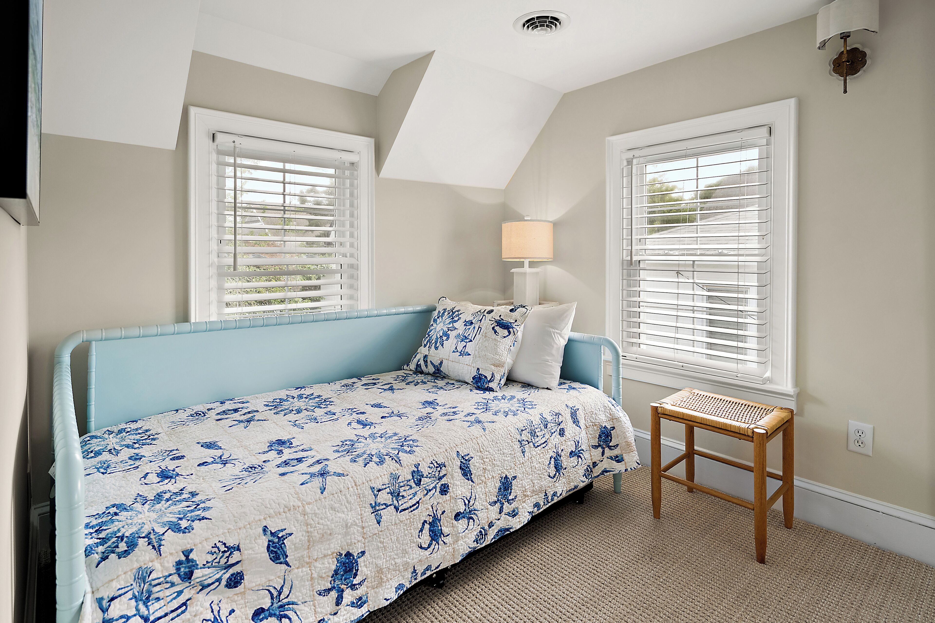 A bedroom with a bed, two windows with blinds, a bedside lamp, and a small wooden stool.