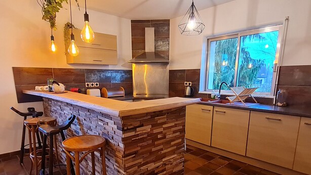A modern kitchen with a central island, bar stools, a window with a view, and hanging pendant lights. - Renovated eco-village cottage, air-conditioned, pet-friendly (Ville-sur-Yron)