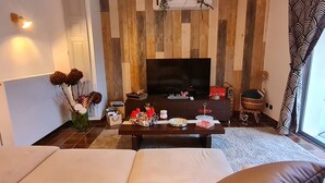 A living room with a flat-screen TV, a wooden accent wall, a sofa, a coffee table with decorative items, and a window with patterned curtains. - Renovated eco-village cottage, air-conditioned, pet-friendly (Ville-sur-Yron)