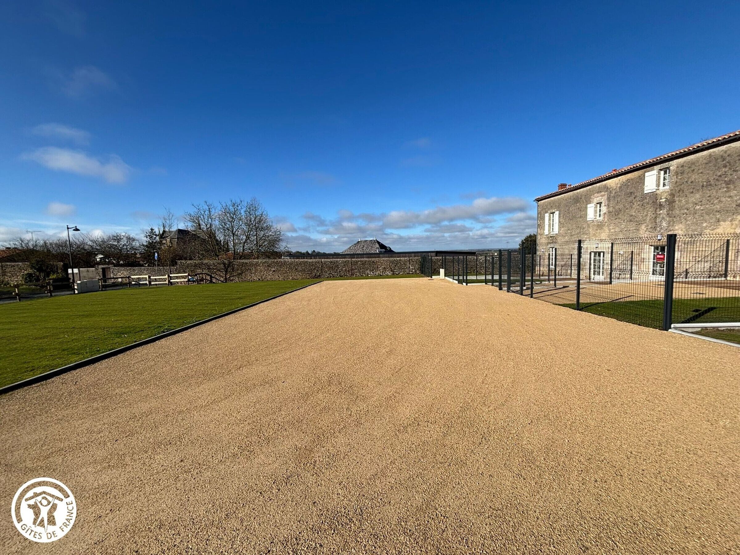 A large indoor sports court with a gravel surface, surrounded by a fence, and a building with a stone facade in the background.