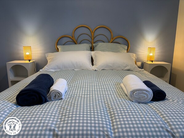 Cama bem arrumada com colcha xadrez, duas luminárias de cabeceira e toalhas enroladas sobre a cama.