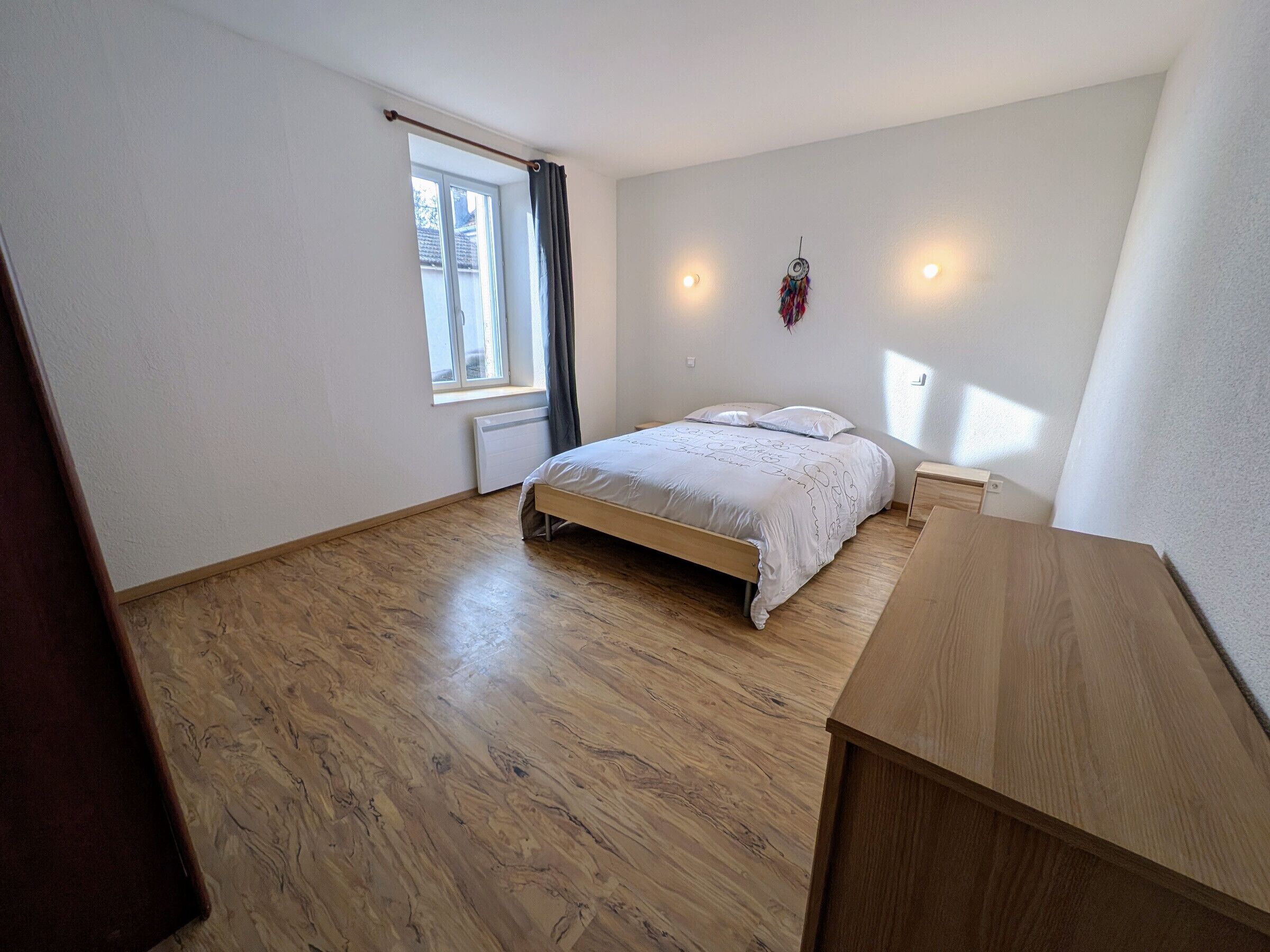 A bedroom with a wooden floor, a bed with a white bedspread, a window with curtains, and a wall-mounted light fixture.