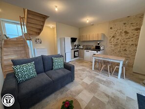 A compact living space with a sofa, a dining table, and a kitchen area including a microwave and a washing machine. - Charming Vendée Cottage with Garden - 6 Guests (La Gaubretière)