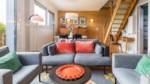 Ein modernes Wohnzimmer mit einer grauen Sofagarnitur, einem Couchtisch mit dekorativer Platte, einem Essbereich mit Holztisch und -stühlen sowie einer Holztreppe.