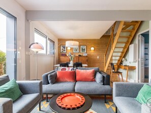 Un salon moderne avec un canapé gris, une table basse agrémentée d’un plateau décoratif, un coin repas avec une table et des chaises en bois, et un escalier en bois.