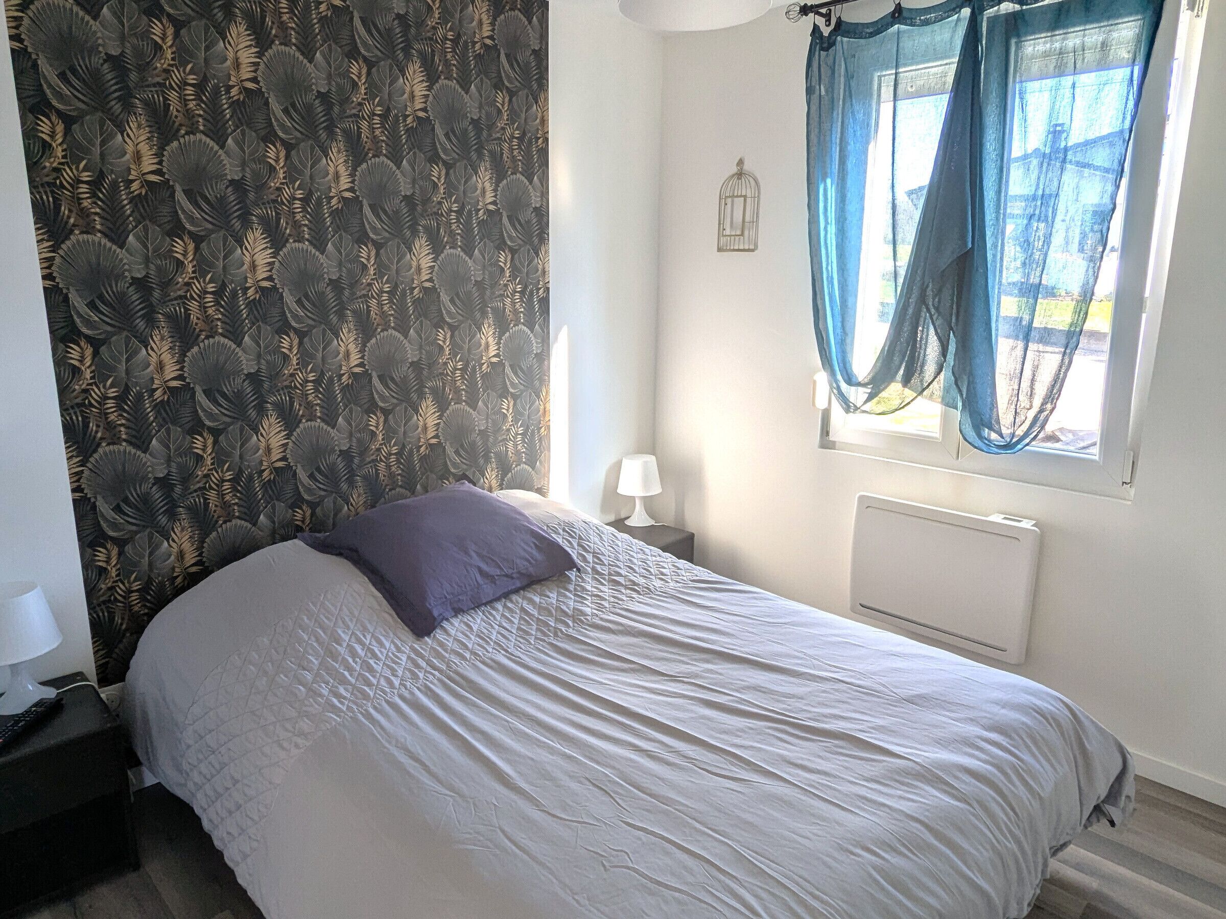 Une chambre à coucher avec un lit, une table de chevet, une lampe, une fenêtre avec des rideaux et un radiateur.