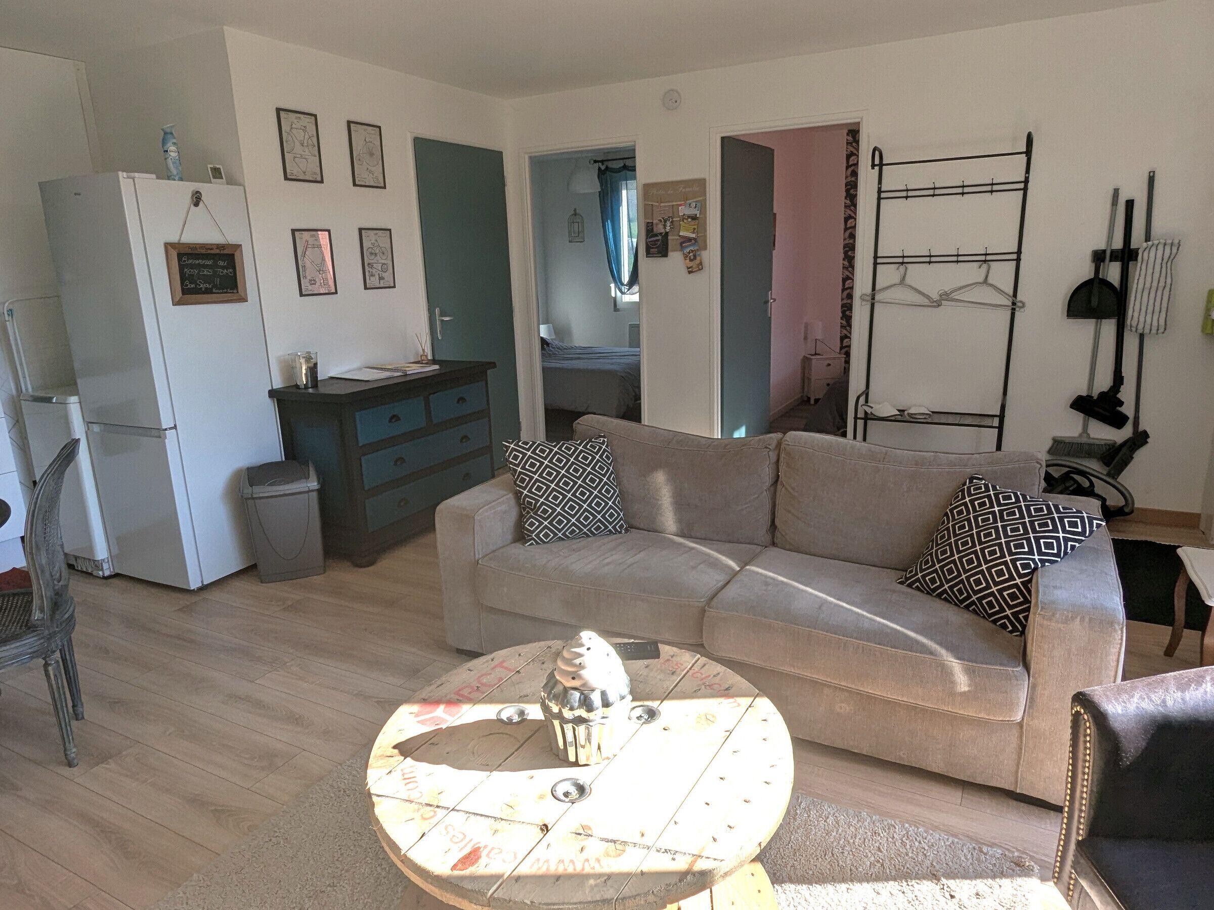 A living room with a sofa, a coffee table, and a refrigerator. There are framed pictures on the wall and a small cabinet.