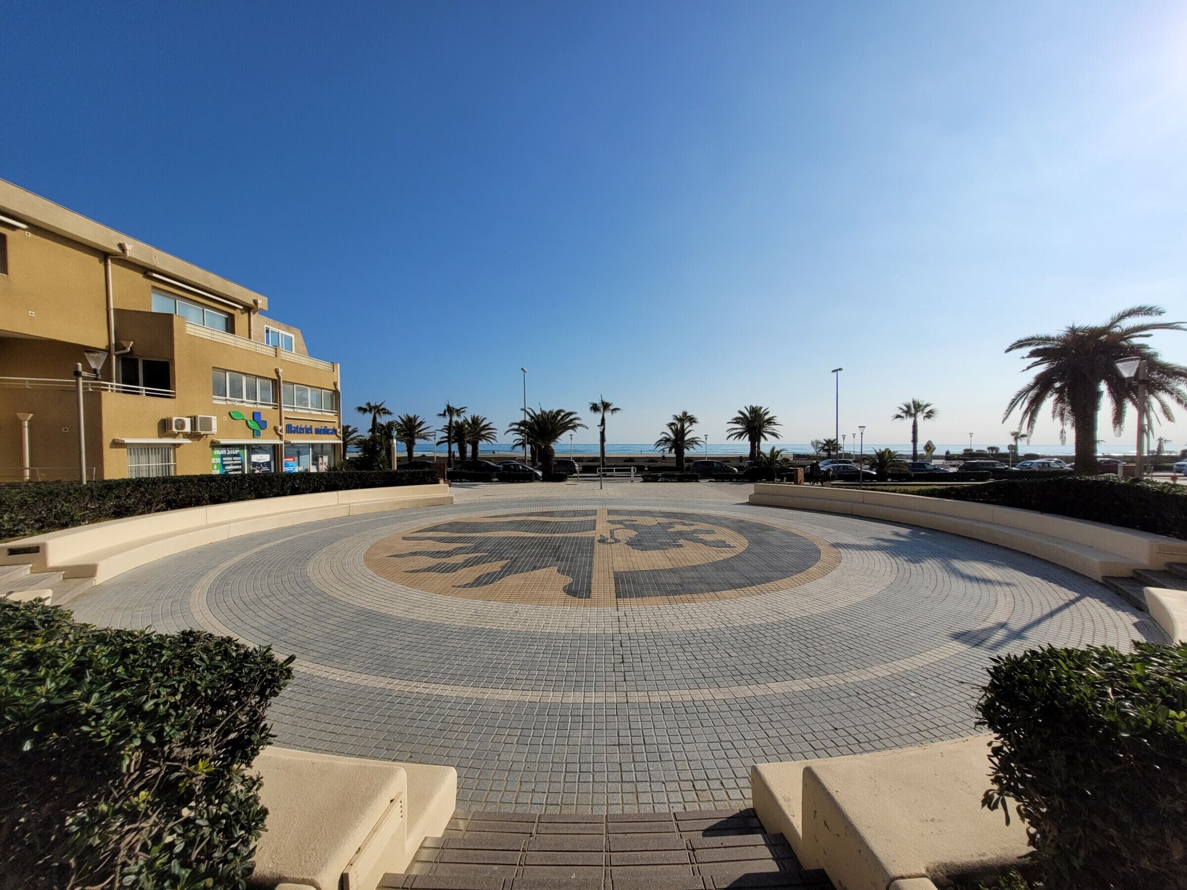 Une place circulaire, agrémentée d’un motif central, de palmiers et d’un bâtiment sur lequel on peut lire l’inscription « Hôtel Merceria ».