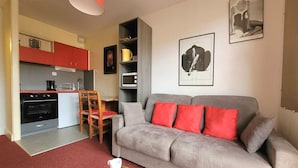 A compact living space with a kitchenette, a grey sofa with red cushions, a dining area with wooden chairs, and a coffee table with cups. - Canet-en-Roussillon: Studio for 2 with balcony (Canet-en-Roussillon)