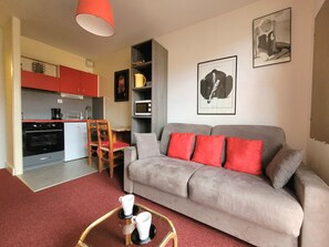A compact living space with a kitchenette, a grey sofa with red cushions, and a dining area with wooden chairs. - Canet-en-Roussillon: Studio for 2 with balcony (Canet-en-Roussillon)