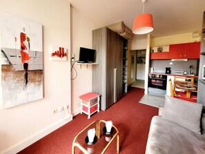 A modern living room with a sofa, a coffee table, and a TV. There is a kitchen area with red cabinets and a dining table. - Canet-en-Roussillon: Studio for 2 with balcony (Canet-en-Roussillon)