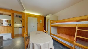 Ein kleines Zimmer mit Etagenbett, Esstisch mit Stühlen, Küchenzeile und einer Tür zu einem weiteren Raum.