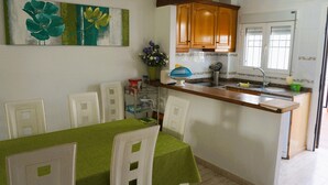Una cucina compatta con zona pranzo, con un tavolo verde, sedie bianche, pensili in legno e un grande quadro di fiori sulla parete.