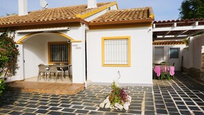 Una casa a due piani con tetto in tegole, un patio con tavolo e sedie, e un piccolo giardino con rocce e piante.