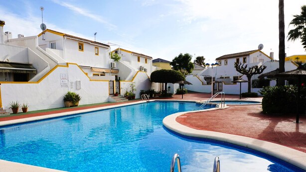 Una piscina con zona solarium rossa, circondata da edifici bianchi con decorazioni gialle e antenne paraboliche.