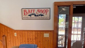 Ein Zimmer mit Holzwandverkleidung, ein Ledersessel mit Kissen und ein Schild mit der Aufschrift 'BAIT SHOP BEER & ICE WORMSMINNOWSTACKLELIES!'.