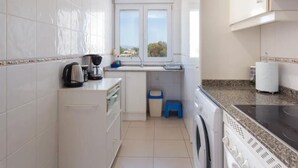 A compact kitchen with white cabinets, a coffee maker, a washing machine, and a window with a view. - Charming 2-Bedroom Apartment in Denia, Beach Access, Parking Included (Denia)